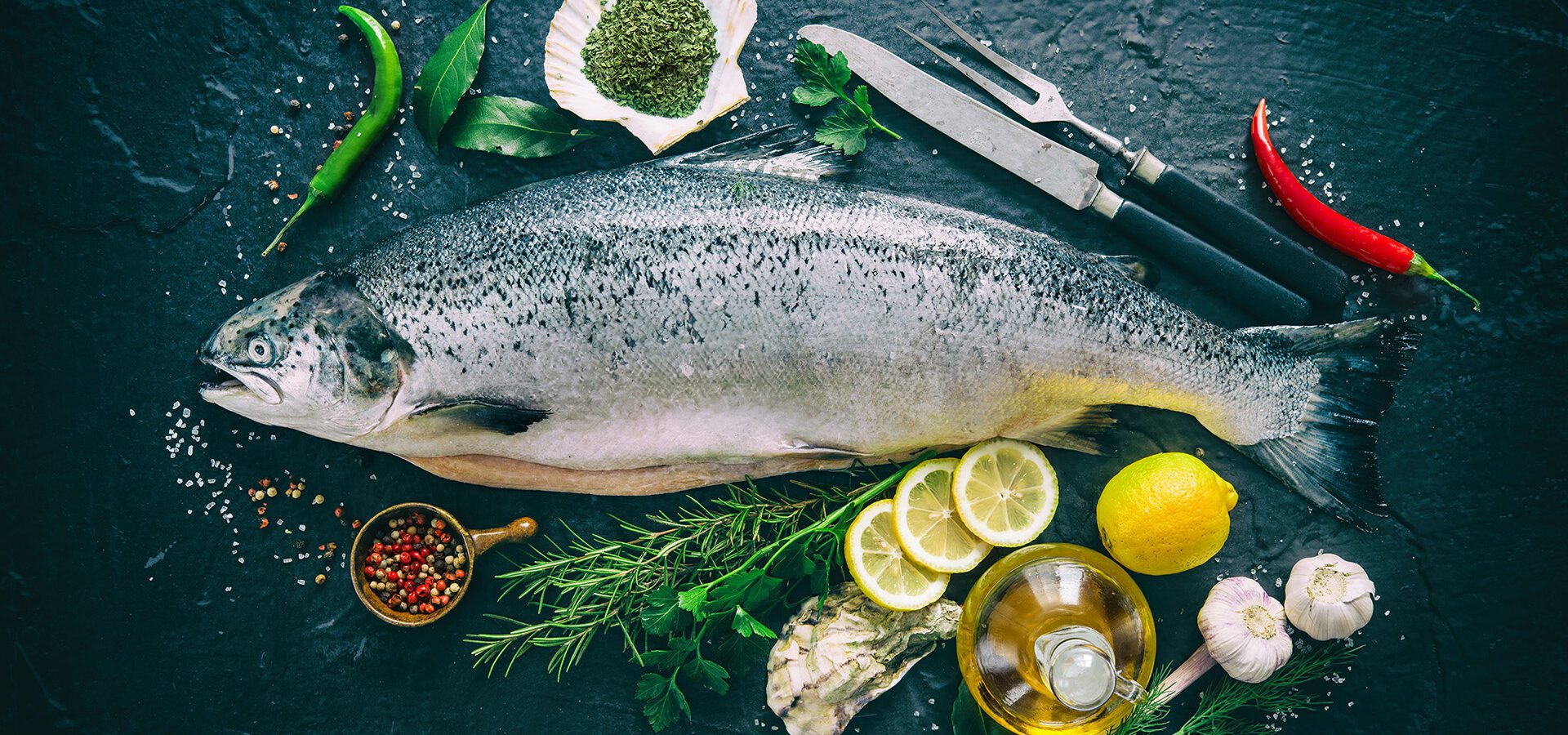 Fresh whole salmon fish with lemons, herbs and seasoning on a rustic black stone background